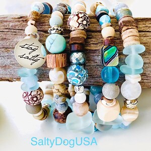 May include: Stack of five bracelets with blue, white, and brown beads. The bracelets feature various textures and patterns, including floral designs, sea glass, and wood. One bracelet has a silver charm with the text "Stay Salty".