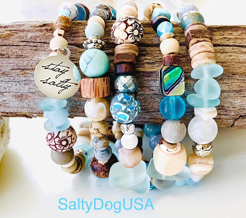 May include: Stack of five bracelets with blue, white, and brown beads. The bracelets feature various textures and patterns, including floral designs, sea glass, and wood. One bracelet has a silver charm with the text "Stay Salty".