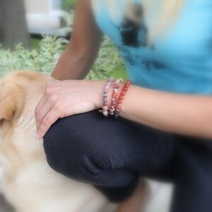 以下が含まれることがあります： 「Live True」と書かれた青いタンクトップを着た女性が犬と一緒に座っています。彼女は手首に3つのブレスレットをしています。1つはピンク、1つは赤、もう1つはピンクと茶色の石が混ざっています。