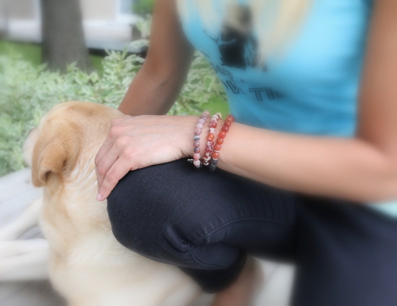 以下が含まれることがあります： 「Live True」と書かれた青いタンクトップを着た女性が犬と一緒に座っています。彼女は手首に3つのブレスレットをしています。1つはピンク、1つは赤、もう1つはピンクと茶色の石が混ざっています。