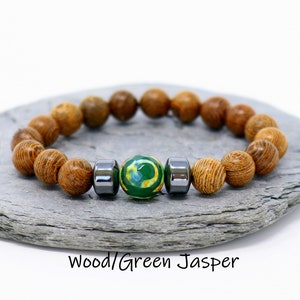 Op de afbeelding: Een houten kralen armband met een groene jaspis steen en twee zwarte hematiet kralen. De armband ligt op een grijs stenen oppervlak. Hout/Groene Jaspis
