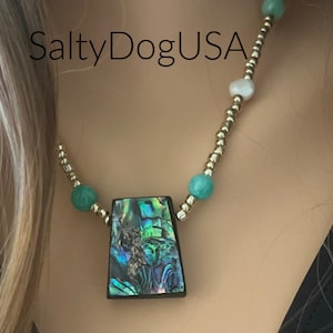 Pode incluir: Um colar de corrente dourada com contas verdes e brancas e um grande pingente de concha de abalone iridescente. O texto "SaltyDogUSA" é visível na imagem.