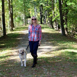 May include: A woman wearing a red and blue plaid shirt, sunglasses, and black boots walks on a path in a forest with a light brown dog on a leash.