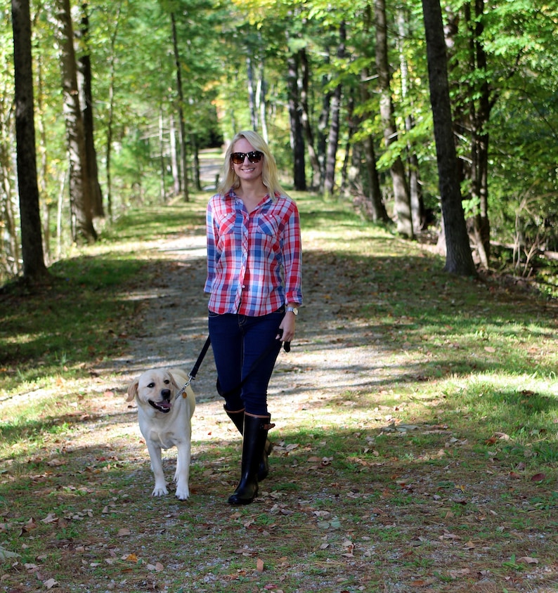 May include: A woman wearing a red and blue plaid shirt, sunglasses, and black boots walks on a path in a forest with a light brown dog on a leash.