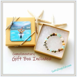 May include: A light blue and brown beaded bracelet with a silver anchor charm. The bracelet is in a small, brown gift box with a white interior. The box is tied with a silver ribbon. The box has a label that says "SaltyDogUSA.com" and "Handmade Unisex Jewelry". The text "Complimentary Gift Box Included" is written below the box.