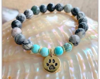 May include: A bracelet made of gray and white marbled stones with turquoise accents and a gold paw print charm.