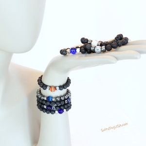 Op de afbeelding: Een set van vijf armbanden met zwarte lavastenen, zilveren kralen en kleurrijke accentkralen. De armbanden zijn gestapeld op de arm van een mannequin. De tekst "SaltyDogUSA.com" is zichtbaar in de rechterbenedenhoek van de afbeelding.