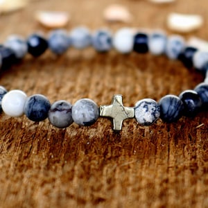 May include: A gray and black beaded bracelet with a silver cross charm. The bracelet is made of natural stones and is a stylish accessory for any occasion.