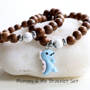 Könnte beinhalten: Ein Set aus zwei Holz-Armbändern mit weißen Perlen und einem blauen Dinosaurier-Anhänger. Die Armbänder liegen auf einem weißen Stein mit dem Text "Mommy & Me Bracelet Set".