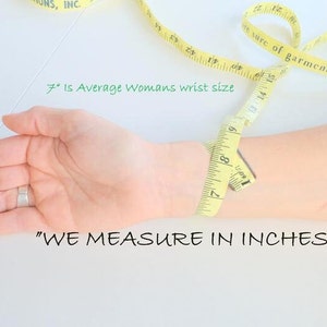 May include: A yellow measuring tape wrapped around a person's wrist. The tape reads "7" inches, which is the average woman's wrist size. The text "WE MEASURE IN INCHES" is written below the wrist.