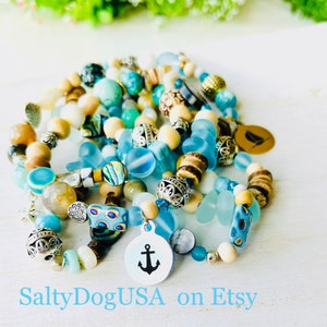 May include: A stack of colorful beaded bracelets with a nautical theme. The bracelets feature blue, white, and brown beads, as well as a silver charm with an anchor. The bracelets are stacked on a white surface. SaltyDogUSA on Etsy.