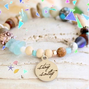 May include: A beaded bracelet with a silver charm that says "stay salty". The bracelet features a variety of beads, including wood, glass, and stone. The beads are in shades of brown, blue, white, and beige.