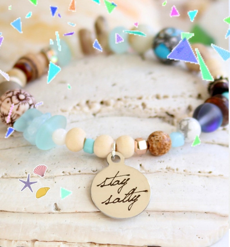 May include: A beaded bracelet with a silver charm that says "stay salty". The bracelet features a variety of beads, including wood, glass, and stone. The beads are in shades of brown, blue, white, and beige.