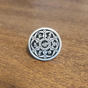 May include: A silver-colored, circular pendant with an intricate design. The pendant features a central floral-like pattern surrounded by six stylized symbols. The background is a warm, wooden surface.