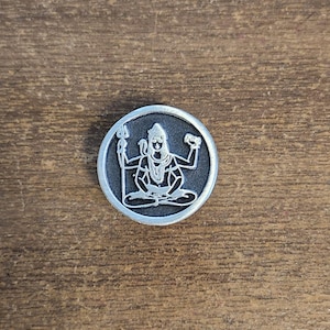 May include: A round, silver-colored metal pendant featuring a detailed depiction of a seated deity with multiple arms, holding various symbolic objects. The deity is surrounded by a circular border. The pendant is set against a wooden background.