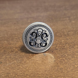 May include: A silver-colored, circular button with a detailed, symmetrical design. The central motif features intertwined shapes, surrounded by a decorative border. The button is likely made of metal and has a slightly raised profile.