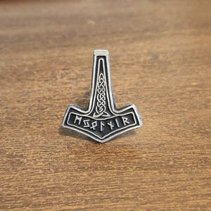 May include: A silver-colored pendant in the shape of Thor's hammer, Mjolnir. The pendant features intricate knotwork and runes. The hammer's head has the word "Mjolnir" inscribed in runes. The pendant is a symbol of Norse mythology.