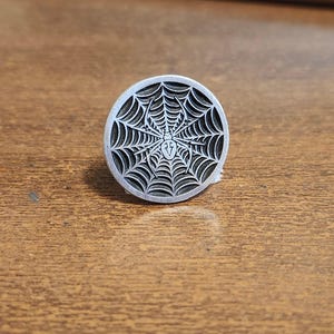 May include: A silver-colored, circular ring featuring a detailed spiderweb design with a spider in the center. The ring is set against a wooden surface, showcasing the intricate details of the metalwork. The ring is a unique piece of jewelry.