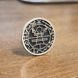 May include: A silver-colored, circular pendant with intricate engraved symbols and patterns. The design features a central figure surrounded by ornate details, creating a detailed and mystical aesthetic. The pendant is likely a piece of jewelry or a decorative item.