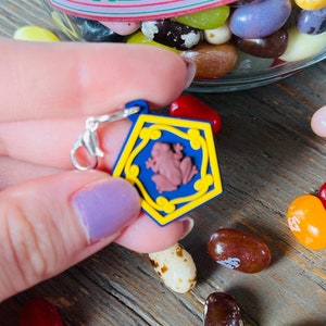 May include: A blue and yellow charm with a frog design, held near a jar of colorful jelly beans. The charm has a silver clasp and a brown frog in the center. The jelly beans are various colors and shapes.