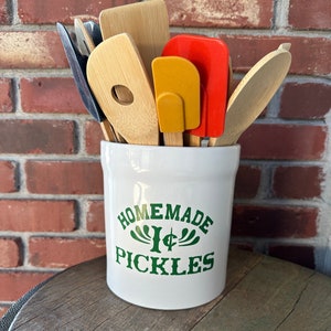 May include: White ceramic utensil holder with green text that reads "Homemade Pickles". The holder is filled with various wooden and silicone spatulas.