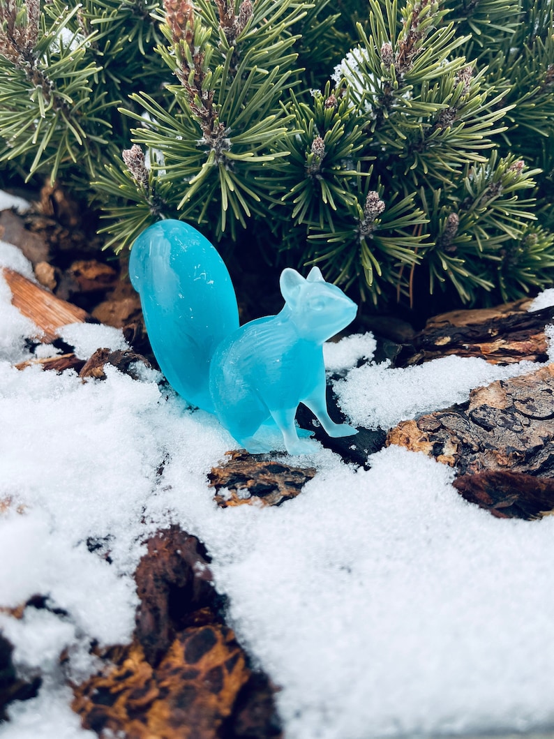 May include: A light blue glass squirrel figurine sitting on a snowy, brown, textured surface.