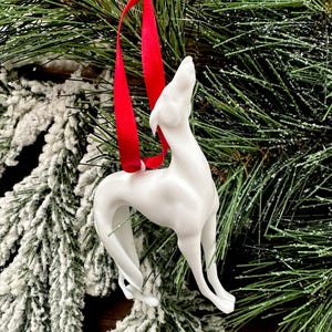 Puede incluir: Adorno de perro galgo blanco de cerámica con una cinta roja para colgar. El perro está posando con la cabeza levantada. El adorno se exhibe sobre un fondo de ramas de pino verdes y nieve artificial.