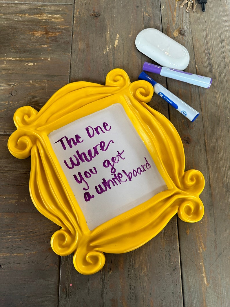 Pu&ograve; includere: Una cornice gialla a tema Friends con un inserto di lavagna bianca cancellabile a secco. La lavagna ha una scritta viola che dice "The One Where You Get A Whiteboard".