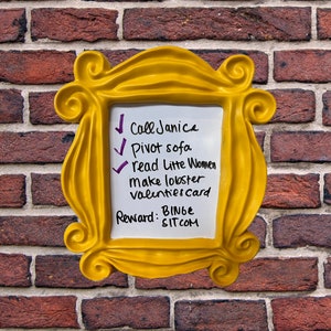 Pu&ograve; includere: Una cornice gialla con una lavagna bianca cancellabile a secco all'interno. La lavagna ha un elenco di compiti scritti su di essa, tra cui "Chiama Janice", "Gira il divano", "Leggi Piccole donne", "Fai l'aragosta", "Biglietto di San Valentino" e "Ricompensa: Binge Sitcom".
