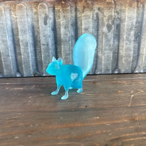 May include: A light blue glass squirrel figurine with a long tail. The figurine is standing on a wooden surface.