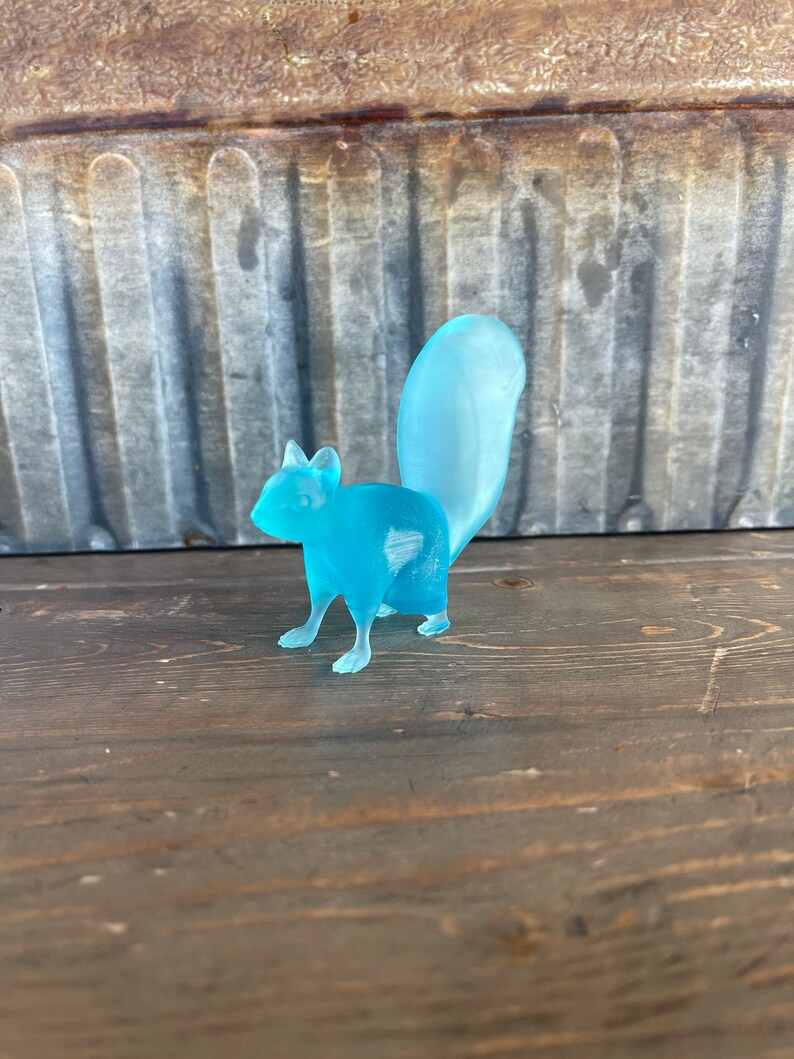 May include: A light blue glass squirrel figurine with a long tail. The figurine is standing on a wooden surface.