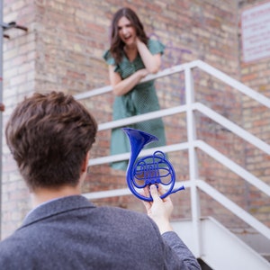 Puede incluir: Una persona sosteniendo un cuerno franc&eacute;s de juguete azul frente a una pared de ladrillo. Una mujer est&aacute; de pie en un tramo de escaleras en el fondo.