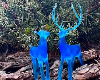 May include: Two translucent blue deer figurines with detailed antlers and slender legs stand on a bed of brown mulch. The deer are in front of a backdrop of green pine needles, creating a striking contrast of colors and textures.