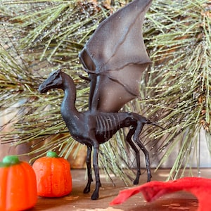 Può includere: Figurina di creatura alata nera con testa di cavallo e corpo scheletrico. La figurina è in piedi su una superficie di legno con due zucche arancioni in primo piano.