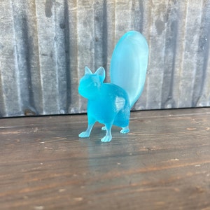 May include: A light blue glass squirrel figurine with a long tail. The figurine is standing on a wooden surface.