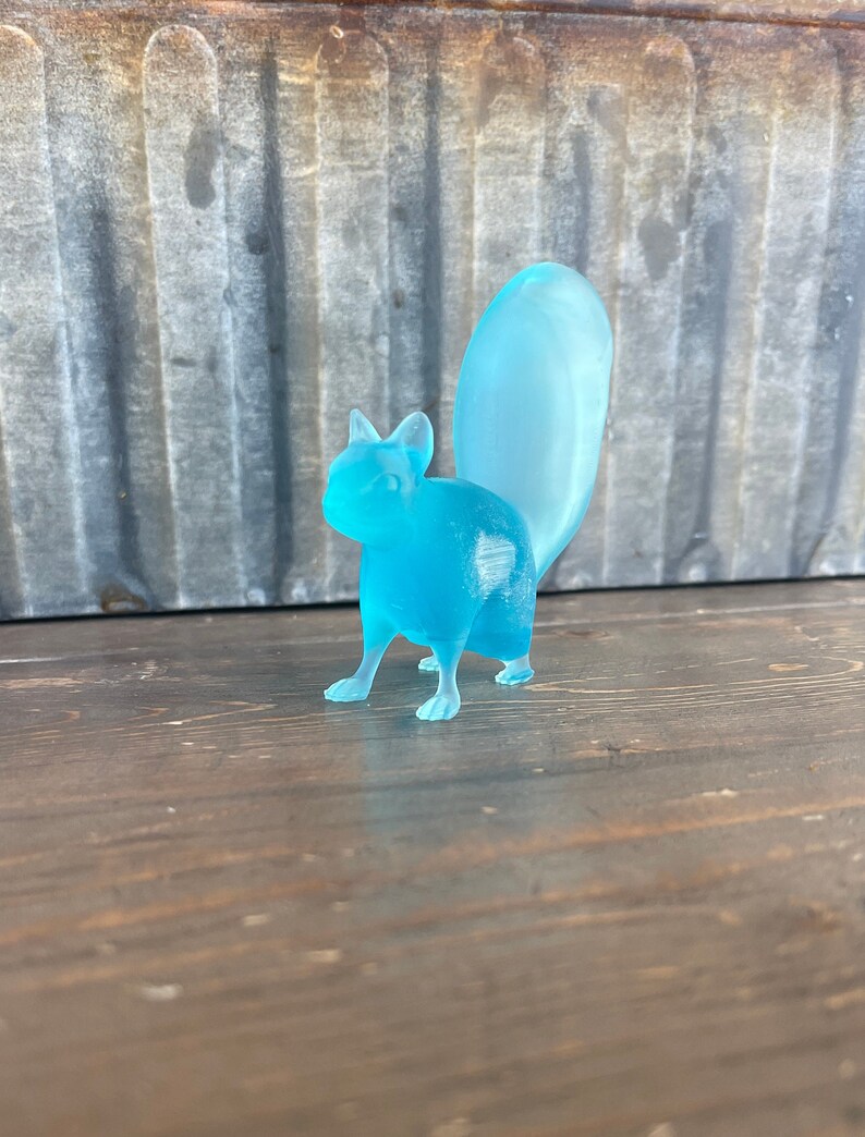 May include: A light blue glass squirrel figurine with a long tail. The figurine is standing on a wooden surface.
