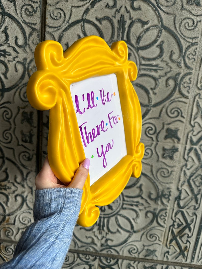 Pu&ograve; includere: Cornice in plastica gialla con un inserto di lavagna bianca cancellabile a secco. La lavagna ha una scritta viola che dice "Sar&ograve; l&igrave; per te."