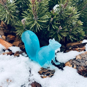 May include: A light blue glass squirrel figurine sitting on a snowy log.