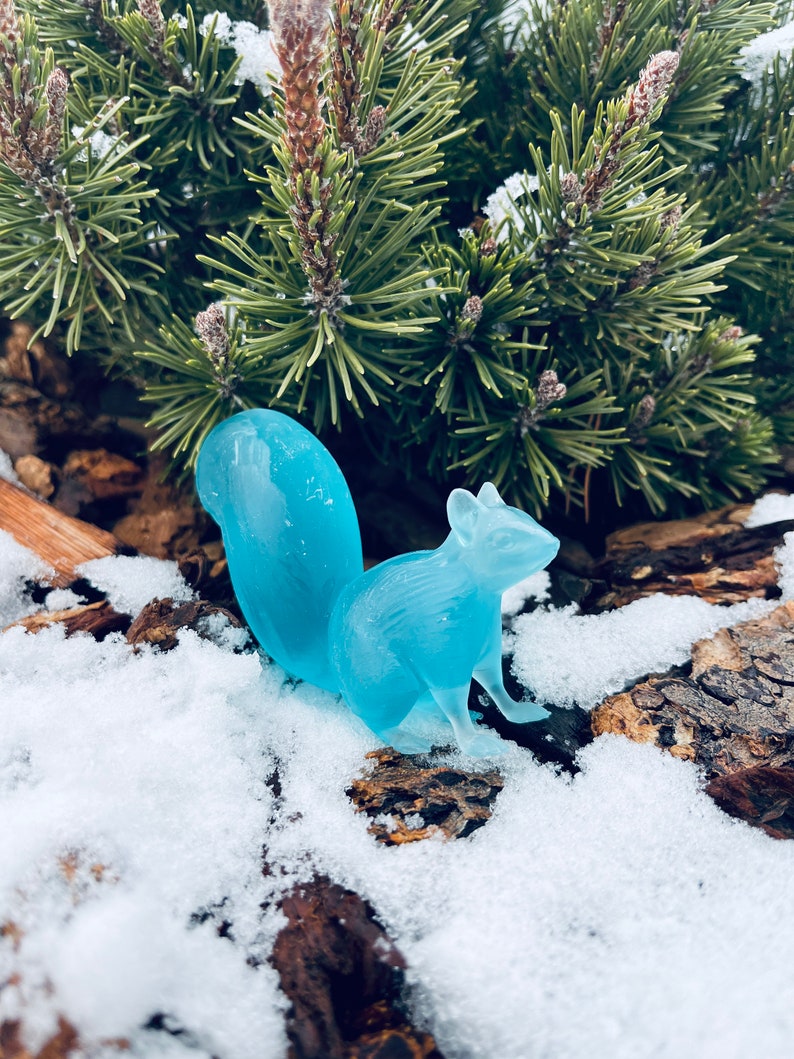May include: A light blue glass squirrel figurine sitting on a snowy log.