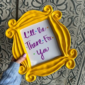 Pu&ograve; includere: Una cornice gialla con sfondo bianco e le parole "I'll be there for you" scritte in viola. La cornice ha la forma di una cornice per foto dello show televisivo Friends.