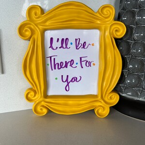 Pu&ograve; includere: Una cornice gialla con sfondo bianco e le parole "I'll be there for you" scritte con un pennarello viola.