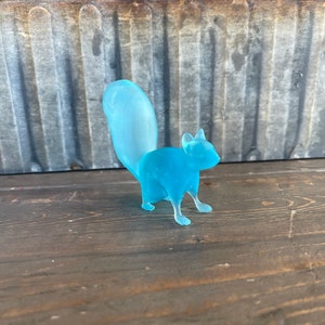 May include: A light blue glass squirrel figurine. The squirrel is standing on a wooden surface.