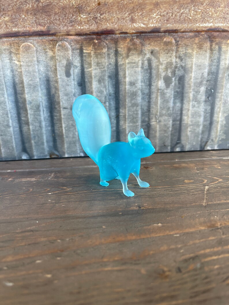 May include: A light blue glass squirrel figurine. The squirrel is standing on a wooden surface.