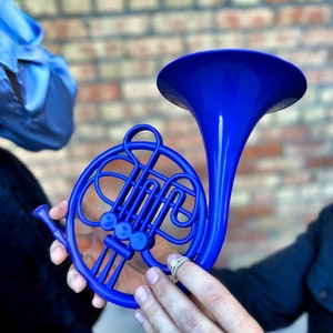 Puede incluir: Un juguete de pl&aacute;stico en forma de trompa azul brillante. La trompa est&aacute; en manos de una persona que lleva un anillo.
