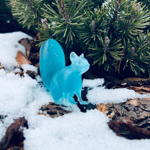 May include: A light blue glass squirrel figurine sitting in the snow. The squirrel has a long tail and is posed in a sitting position.