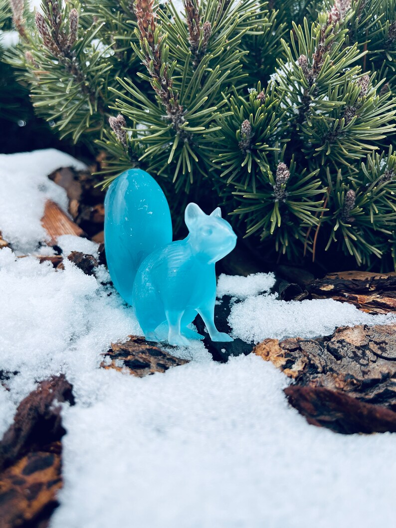 May include: A light blue glass squirrel figurine sitting in the snow. The squirrel has a long tail and is posed in a sitting position.