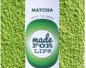 May include: A white tube of matcha lip balm with green accents. The tube says "MATCHA" and "TWIST TO OPEN". It also says "made FOR LIPS" with a heart in the word "FOR". The tube is sitting on a bed of green matcha powder. The net weight is 0.25 oz (7g).