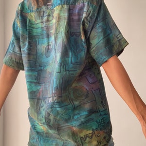 May include: A short-sleeved button-up shirt with an abstract, colorful print in shades of blue, green, brown, and purple. The shirt has a relaxed fit and a collar.