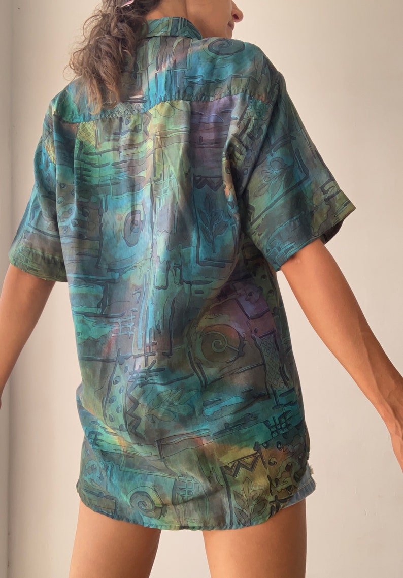 May include: A short-sleeved button-up shirt with an abstract, colorful print in shades of blue, green, brown, and purple. The shirt has a relaxed fit and a collar.