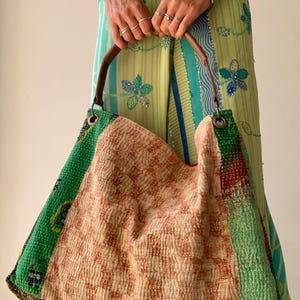 May include: A large patchwork tote bag with a brown leather handle. The bag features a central section of orange and cream quilted fabric, flanked by green patterned fabric. The bag is held by a person wearing a green and blue patterned outfit.